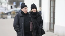 Starszy mężczyzna i kobieta ubrani na czarno spacerują razem po chodniku w chłodny dzień, oboje mają czapki i kurtki, a ich wyraz twarzy jest poważny.