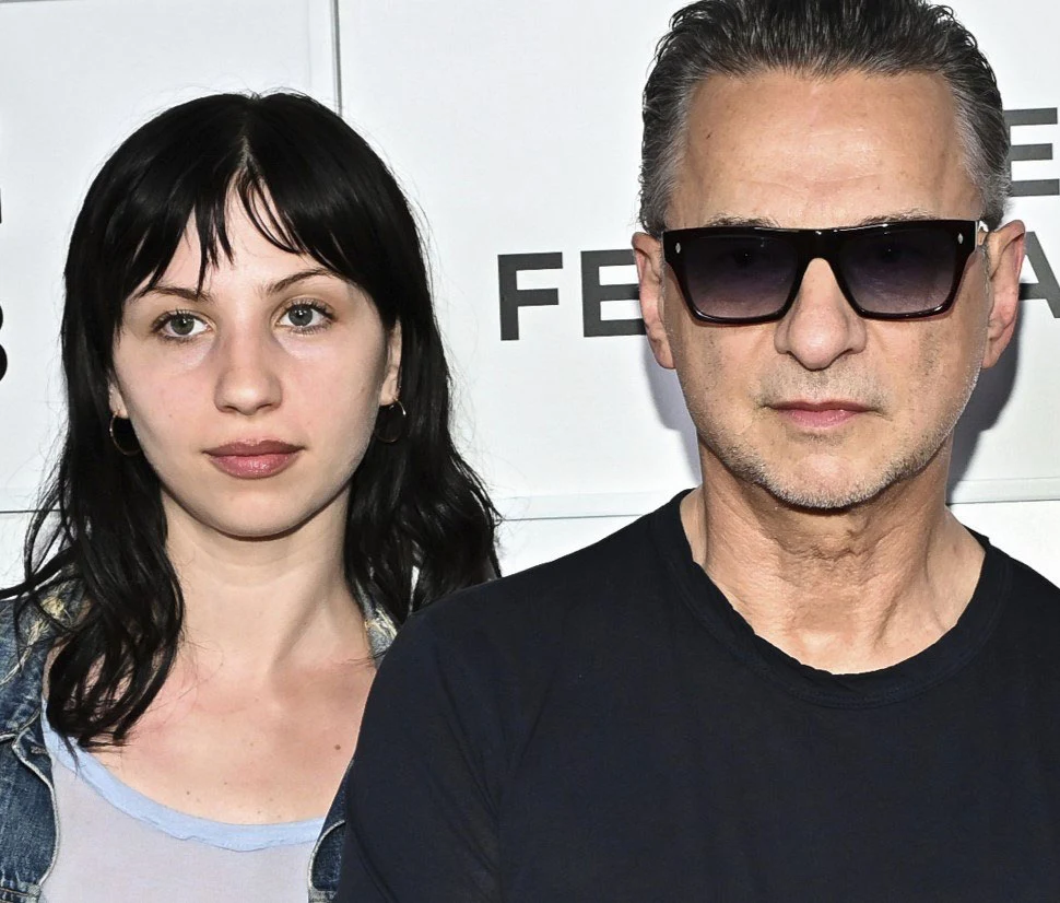 Stella Rose z ojcem Davem Gahanem Dwie osoby stoją obok siebie na tle białej ściany z napisem, jedna z nich nosi ciemne okulary przeciwsłoneczne i czarną koszulkę, druga ma ciemne, proste włosy i ubrana jest w jasną koszulkę oraz jeansową kurtkę.