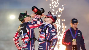 Dwóch sportowców w kombinezonach z flagą Wielkiej Brytanii pije szampana, obok nich trzeci zawodnik trzyma butelkę, w tle jasne fajerwerki.