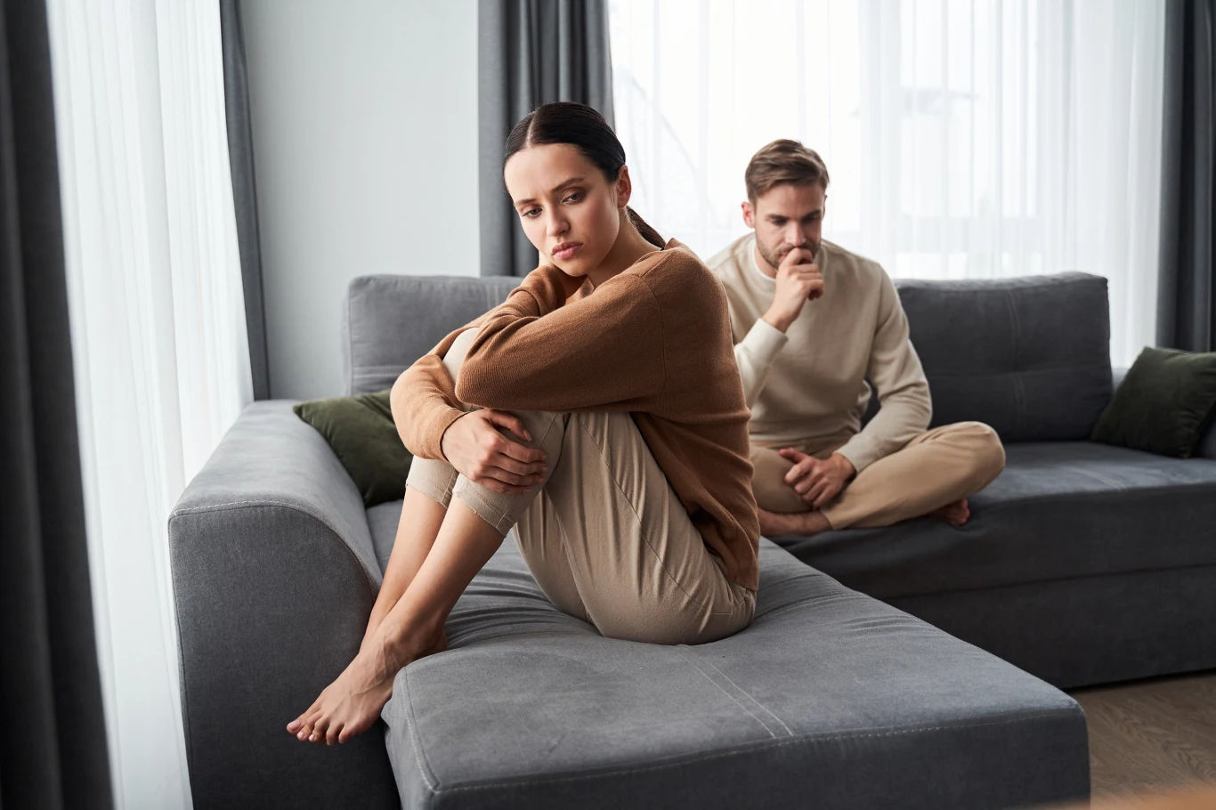 Młoda kobieta siedząca na kanapie ze skrzyżowanymi nogami i smutnym wyrazem twarzy, w tle młody mężczyzna w zamyśleniu, oboje w spokojnym, jasnym salonie.