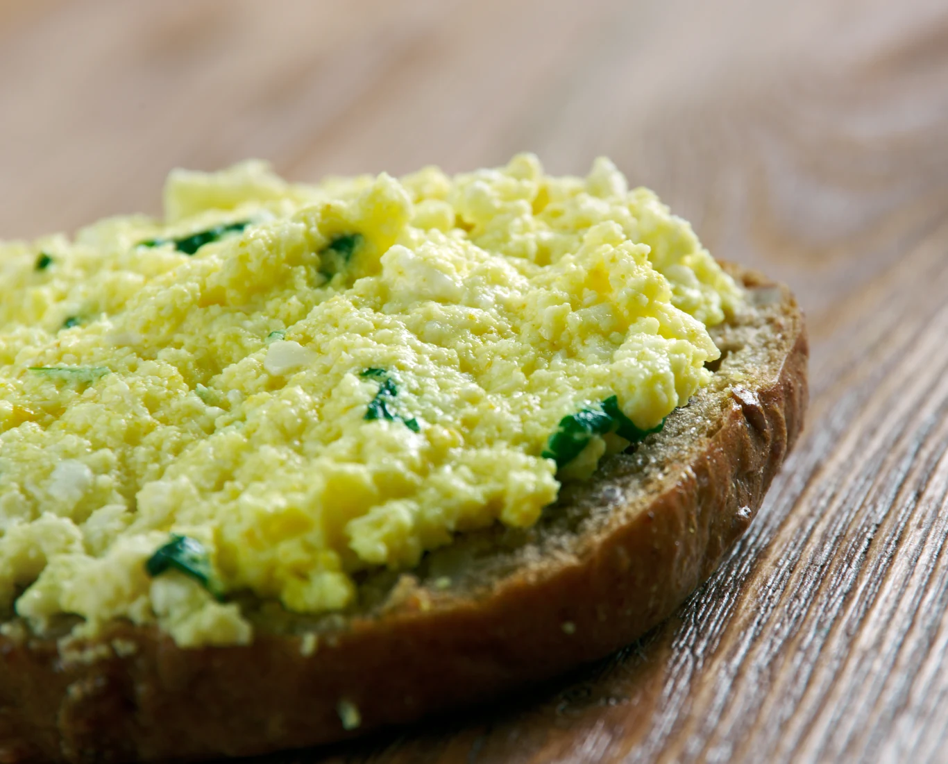 Kromka chleba posmarowana pastą jajeczną z widocznymi kawałkami szczypiorku, ustawiona na drewnianym blacie.