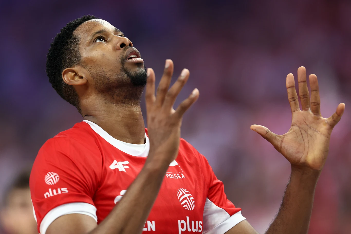 Wilfredo Leon Siatkarz w czerwonej koszulce z białymi napisami i logotypami, patrzący w górę, z uniesionymi dłońmi, stojący na hali sportowej podczas meczu.