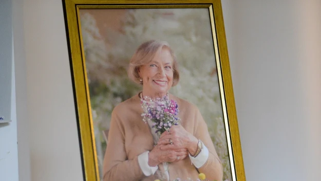 Uśmiechnięta starsza kobieta trzyma bukiet polnych kwiatów, ubrana w jasny sweter, sfotografowana na miękkim tle, obraz umieszczony w złotej ramce.