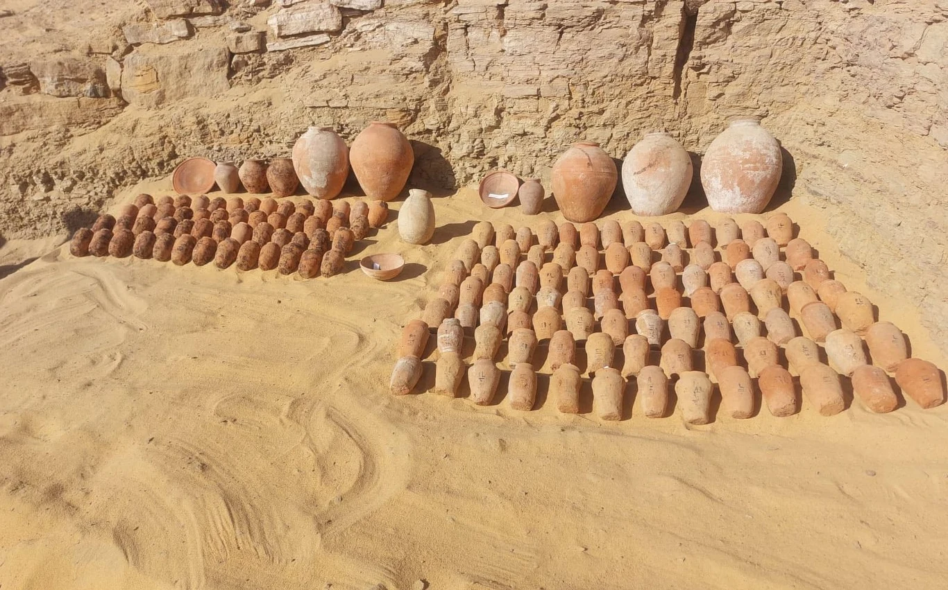 Grobowce i 160 starożytnych naczyń. Wyjątkowe odkrycie w Egipcie Rzędy glinianych naczyń i dzbanów ustawione na piasku w wykopie archeologicznym, w tle widoczny fragment ziemnej ściany.