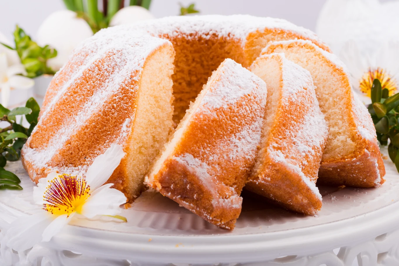 Zwróć uwagę na te techniczne aspekty, a babka pięknie wyrośnie i nie opadnie Pokrojona babka piaskowa posypana cukrem pudrem, ustawiona na białej paterze, udekorowana kwiatami i zielonymi gałązkami.