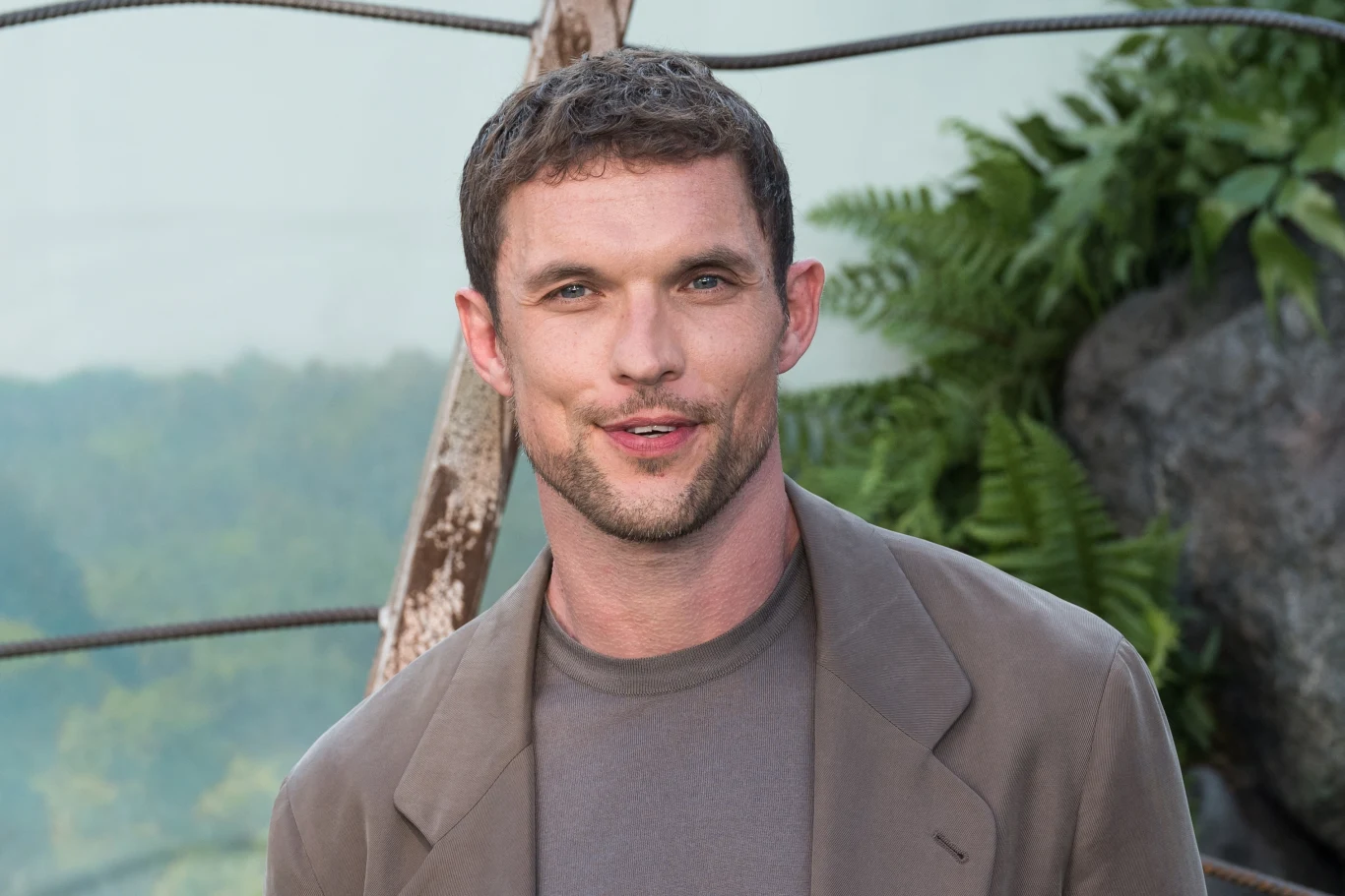 Mężczyzna z krótkimi, brązowymi włosami w szarej marynarce i t-shircie stoi na tle roślin oraz skalnych elementów dekoracyjnych, w naturalnym świetle