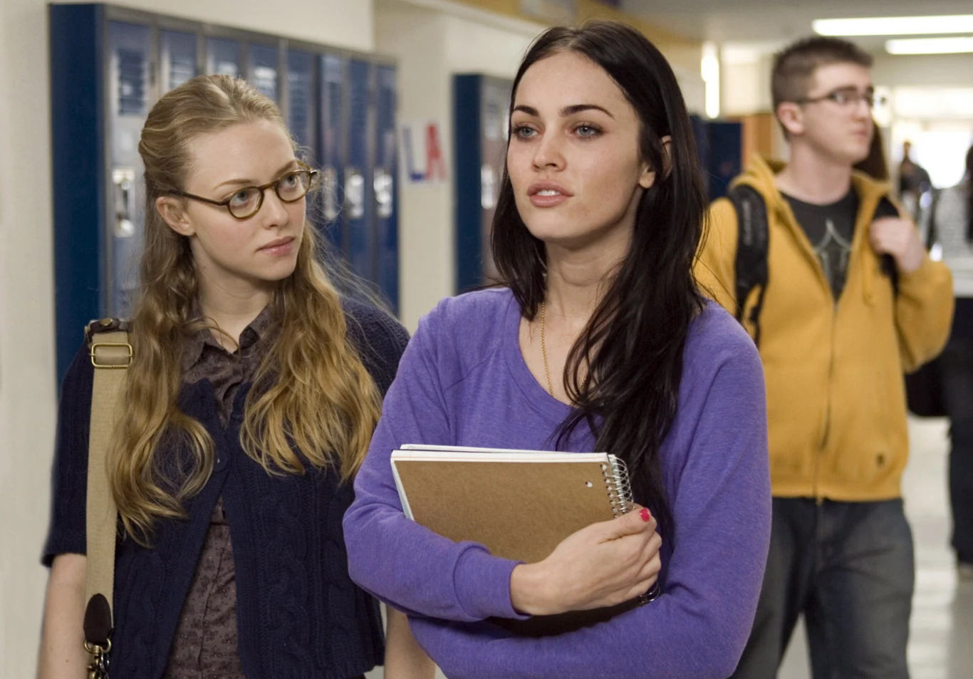 "Zabójcze ciało" Dwie młode kobiety idące szkolnym korytarzem, jedna z nich trzyma zeszyt i patrzy przed siebie, druga spogląda na nią z zaciekawieniem. W tle widać szafki szkolne oraz przechodzących uczniów.