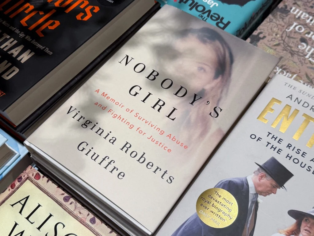 Stos książek ułożonych na półce, z widoczną na pierwszym planie okładką książki 'Nobody's Girl' autorstwa Virginii Roberts Giuffre opowiadającej o przetrwaniu przemocy i walce o sprawiedliwość.
