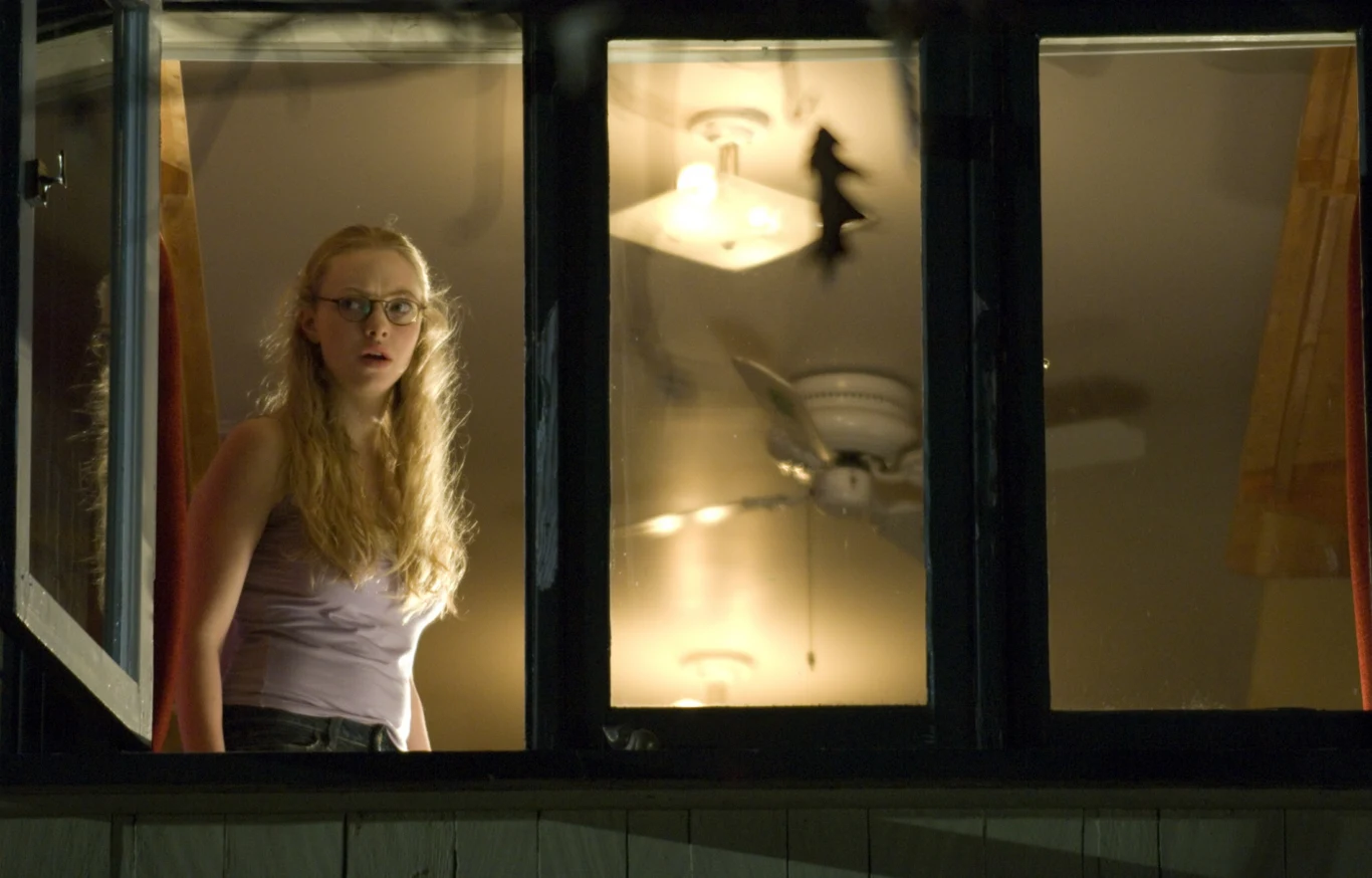 "Zabójcze ciało" Młoda kobieta w okularach patrzy z niepokojem przez otwarte okno, w tle oświetlona lampa sufitowa i wentylator, nad lampą widoczny nietoperz.