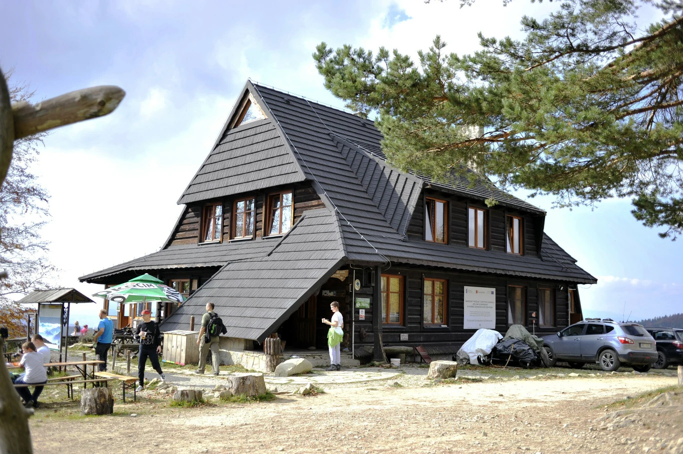 Drewniany górski schronisko z dwuspadowym dachem, otoczone przez ludzi wypoczywających na ławkach i turystów, stojących przed wejściem, w tle widoczne drzewa i zaparkowany samochód.