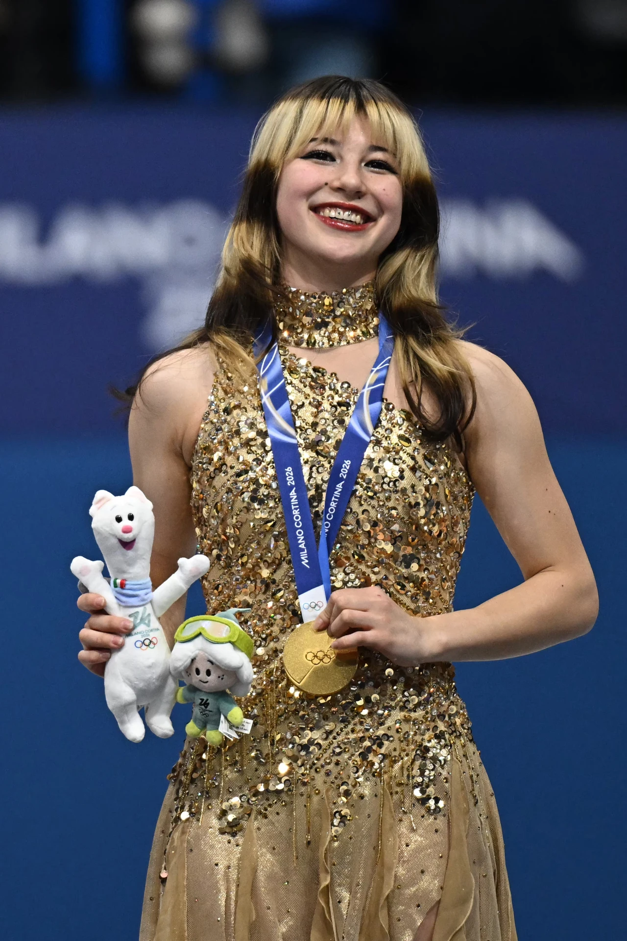 Uśmiechnięta młoda kobieta w złotej sukni trzyma złoty medal olimpijski na niebieskiej wstążce oraz dwie maskotki, tło w kolorze niebieskim z rozmytym tekstem.