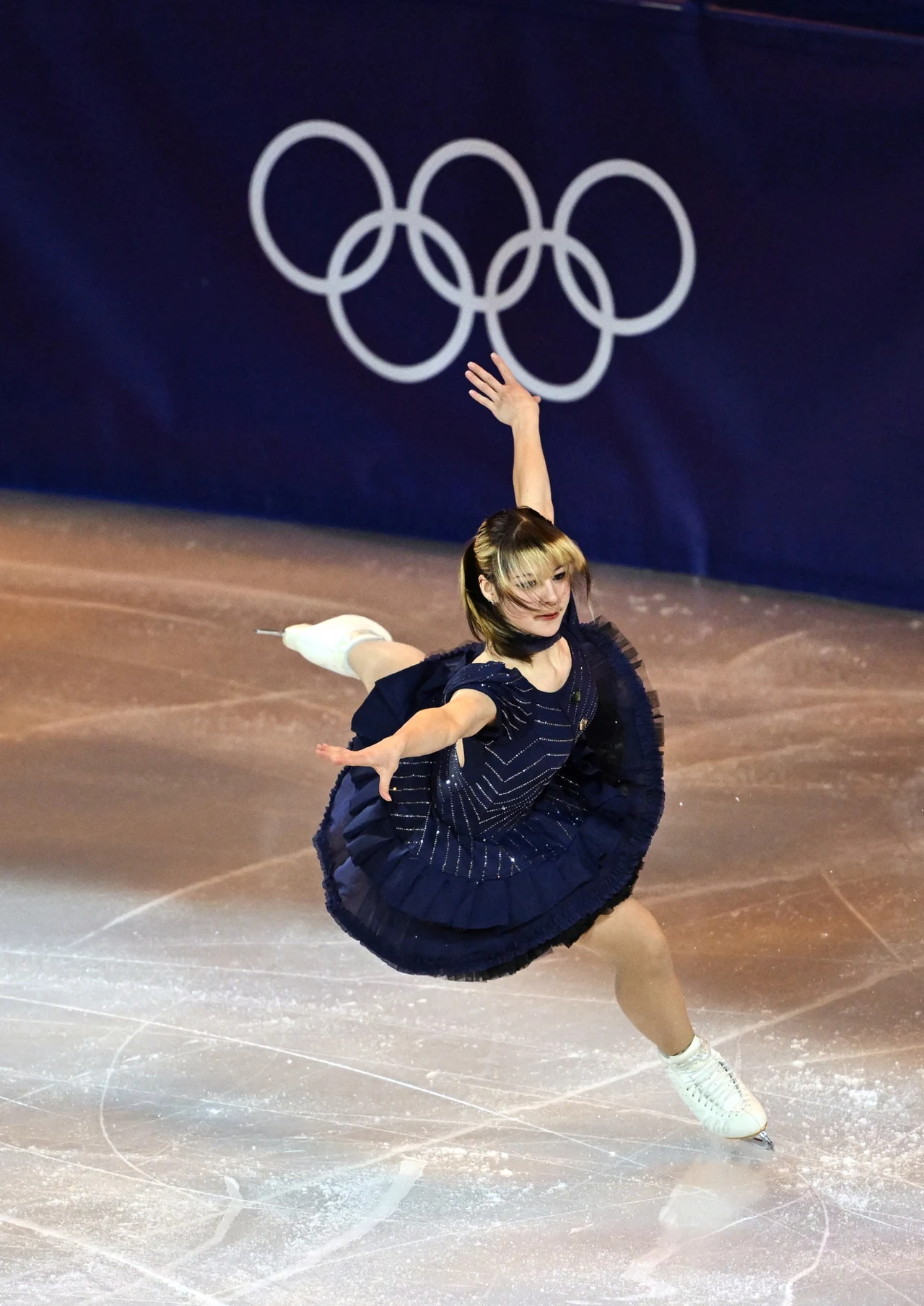 Łyżwiarka figurowa w ciemnej sukni wykonuje efektowny skok na lodowisku, w tle widoczne są olimpijskie koła na granatowej ścianie.