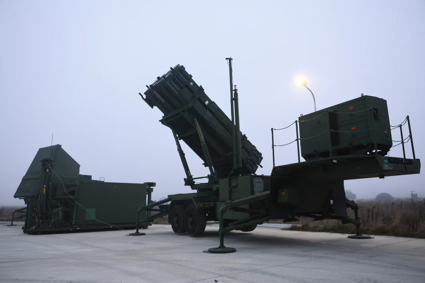 Wojskowy system rakietowy Patriot ustawiony na betonowej nawierzchni na otwartej przestrzeni, w tle lekka mgła i zapalone światło uliczne.
