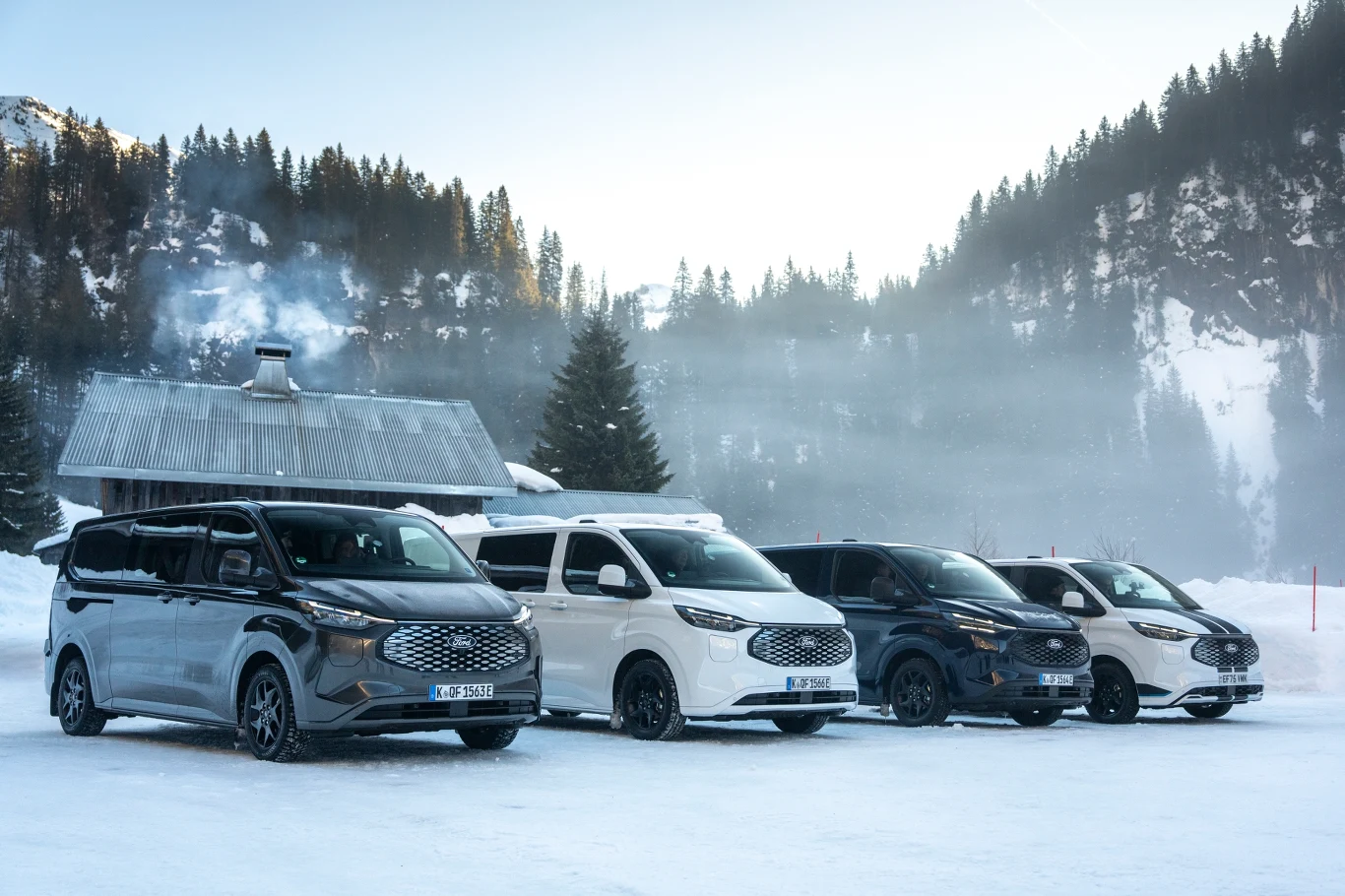 Cztery nowoczesne vany zaparkowane na śniegu przed drewnianym domkiem z kominem, z którego unosi się dym, w tle ośnieżone drzewa i górski krajobraz.