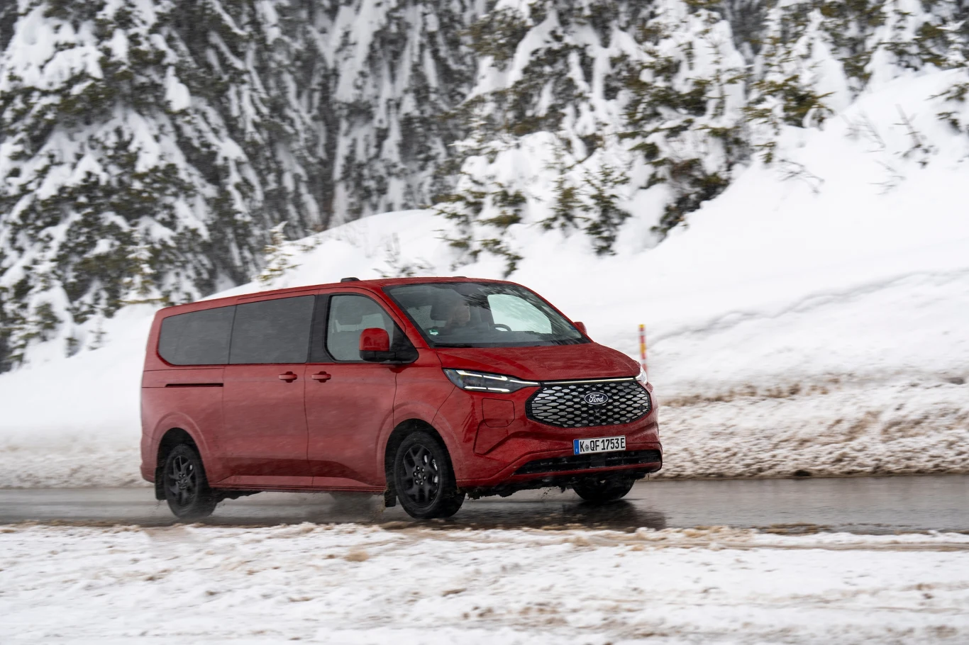 Czerwony minivan jedzie po zaśnieżonej drodze w otoczeniu ośnieżonych drzew i wysokich zasp śnieżnych, koła pojazdu pokrywają się błotem pośniegowym, a w tle widać las.