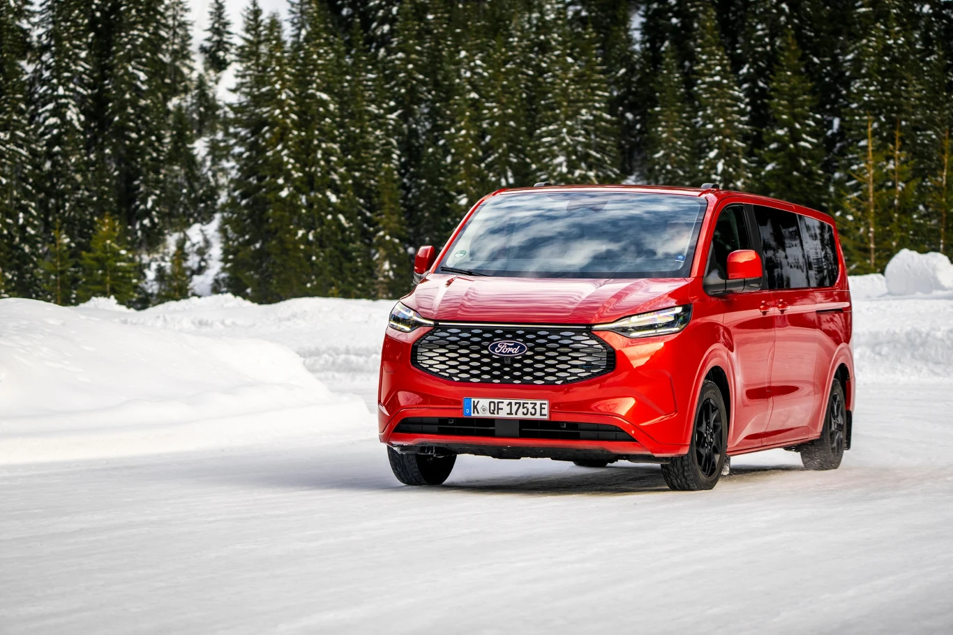 Czerwony samochód osobowy Ford jadący po zaśnieżonej drodze, otoczony wysokimi świerkami w zimowym krajobrazie.