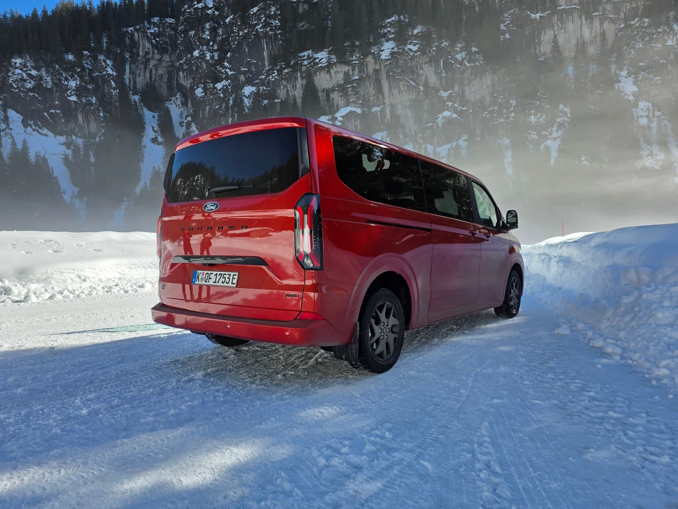 Czerwony van marki Ford zaparkowany na zaśnieżonej drodze, otoczony zimowym krajobrazem z wysokimi ośnieżonymi skałami i drzewami w tle.
