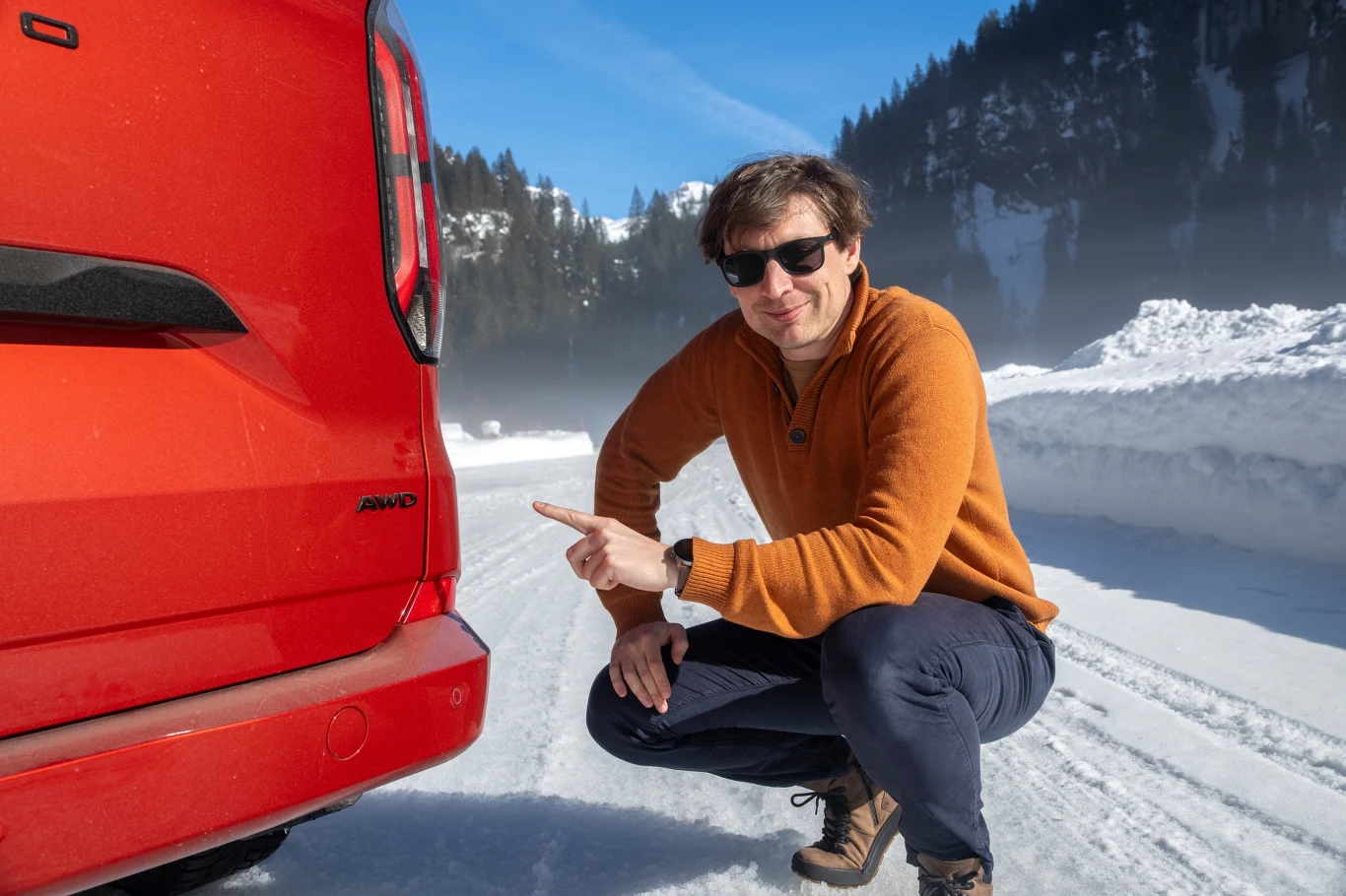 Mężczyzna w okularach przeciwsłonecznych w pomarańczowym swetrze kuca przy czerwonym samochodzie zaparkowanym na zaśnieżonej drodze w górskim otoczeniu, wskazując palcem na emblemat auta.