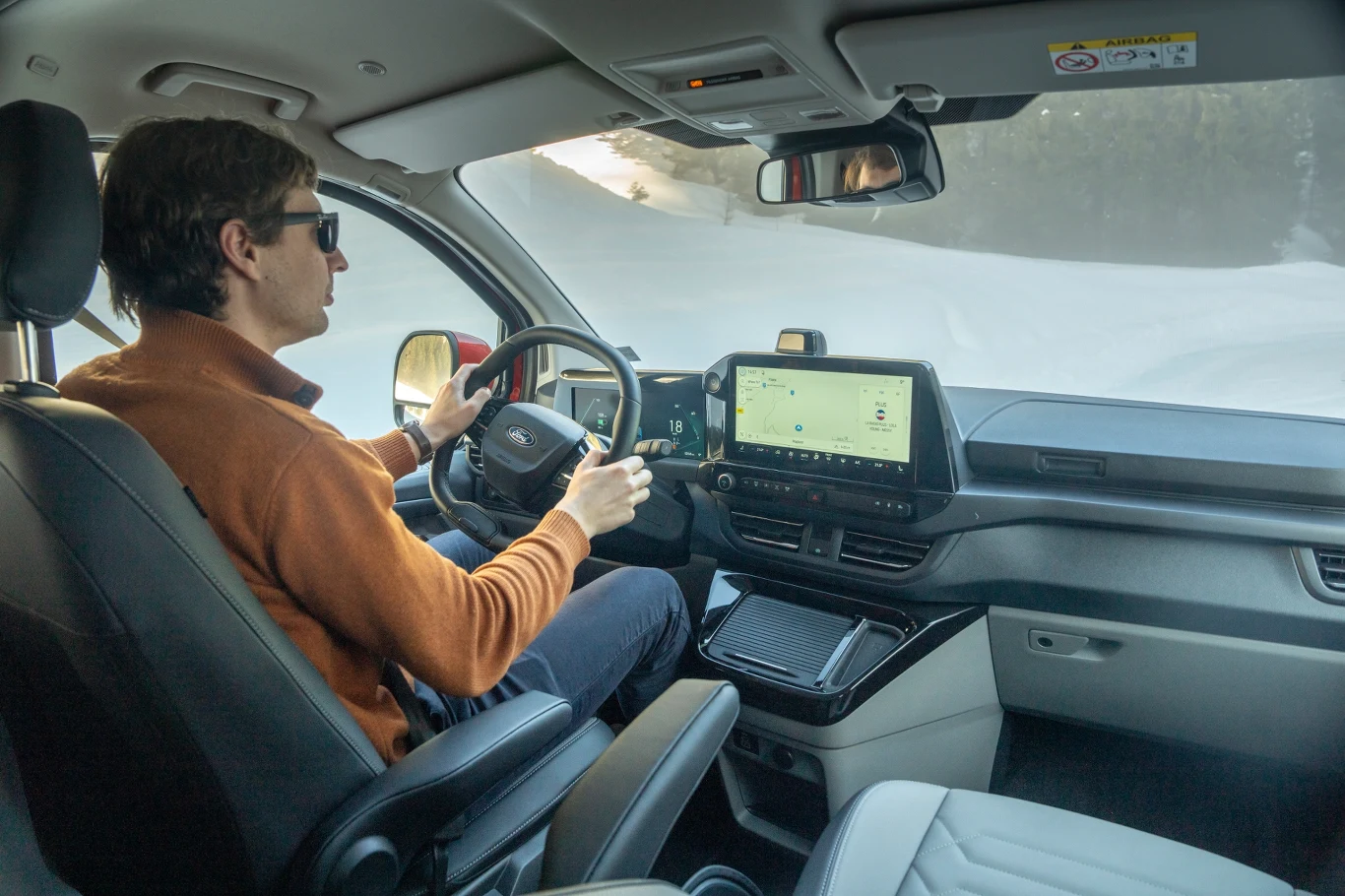 Mężczyzna w okularach przeciwsłonecznych prowadzi nowoczesny samochód, skupiając uwagę na drodze, we wnętrzu pojazdu dominuje centralnie umieszczony ekran dotykowy oraz jasne i przestronne wykończenie kabiny.