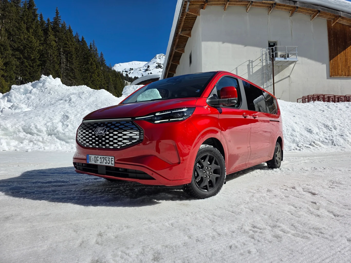 Czerwony minivan Ford Tourneo Custom stoi na zaśnieżonej drodze, w tle białe góry, las iglasty i budynek z drewnianym wykończeniem dachu.