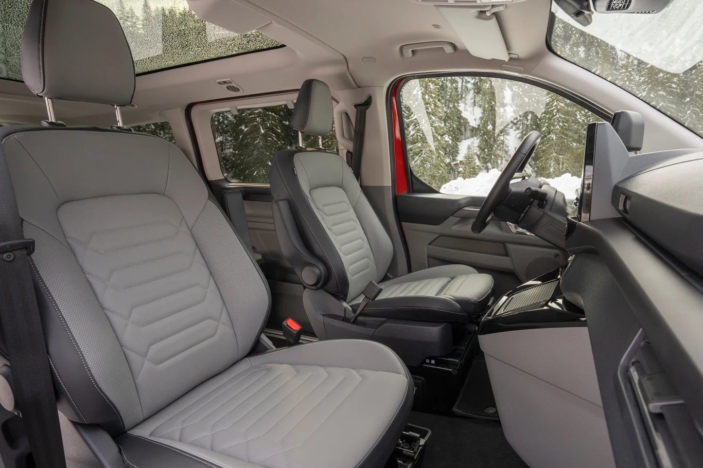 Przestronne wnętrze nowoczesnego samochodu z dwoma szarymi fotelami obitymi skórą, nowoczesną deską rozdzielczą oraz widokiem na zasypany śniegiem krajobraz przez panoramiczny dach i boczne okna.