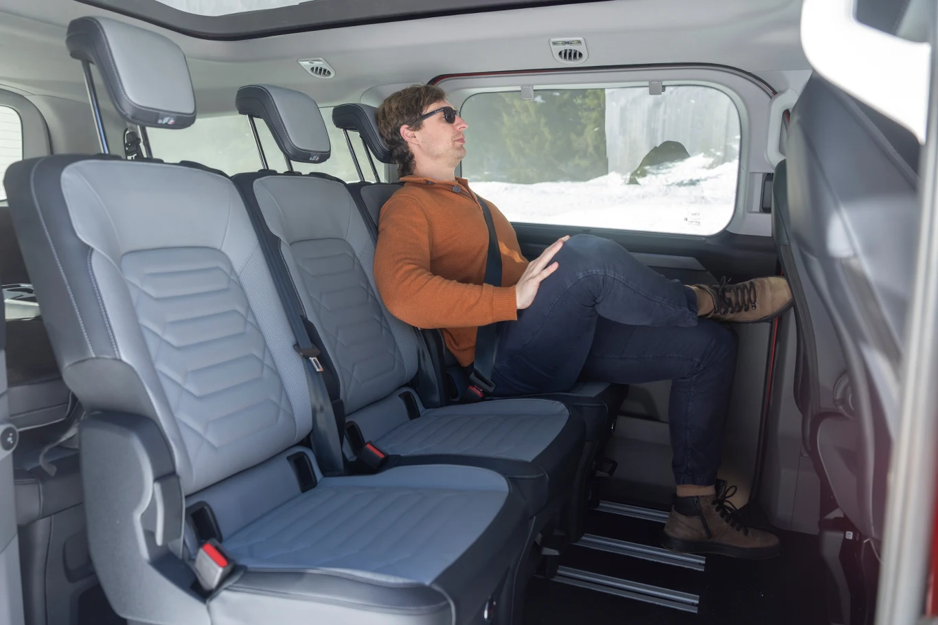 Mężczyzna w okularach przeciwsłonecznych siedzi zrelaksowany na tylnym siedzeniu przestronnego samochodu osobowego, nogi założone na siebie, wnętrze jasne i nowoczesne.