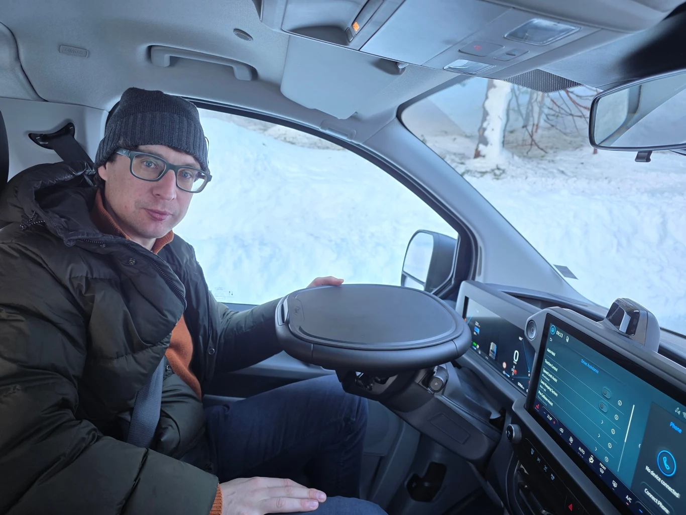 Mężczyzna w okularach i zimowej czapce siedzi w nowoczesnym samochodzie, trzyma ręce na okrągłej, nietypowej kierownicy, a za oknem widoczny jest śnieg.