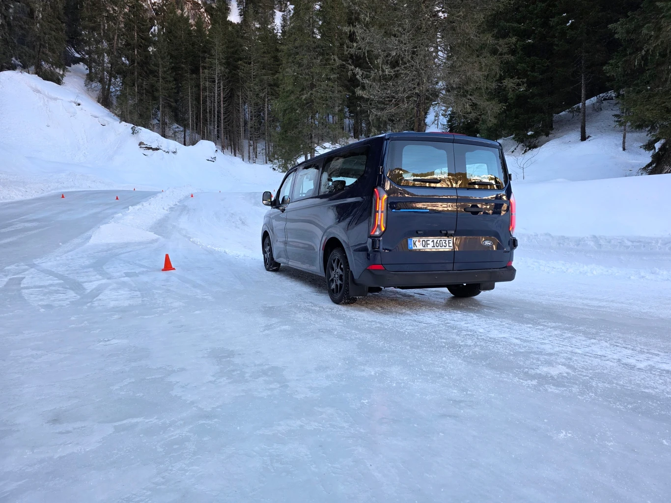 Samochód osobowy jadący po ośnieżonej, oblodzonej drodze leśnej, w otoczeniu drzew iglastych i śnieżnych zasp, z widocznymi pomarańczowymi pachołkami wyznaczającymi trasę.