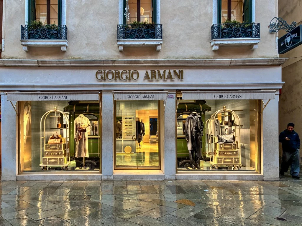 Luksusowa witryna sklepu Giorgio Armani z eleganckimi kreacjami na wystawach, złote światła podkreślają ekskluzywność marki, mokry chodnik i przechodzień dodają scenerii miejskiego charakteru.