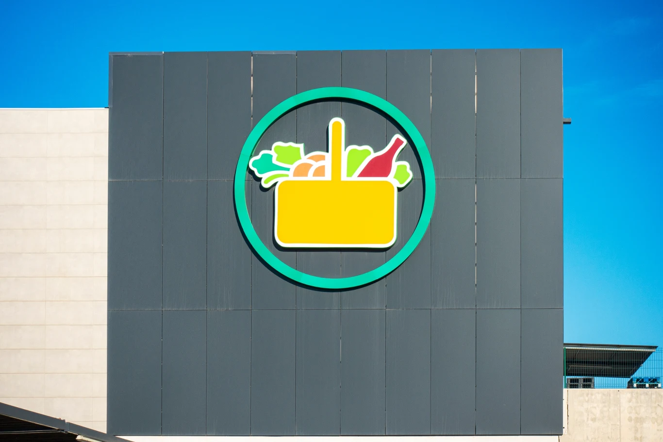 Hiszpańska sieć supermarketów, Mercadona, rozdziela między pracowników miliard euro Logo sklepu spożywczego z żółtym koszykiem na tle okręgu w zielonej ramce, wypełnionym grafikami warzyw i butelki, umieszczone na szarej elewacji budynku pod błękitnym niebem.