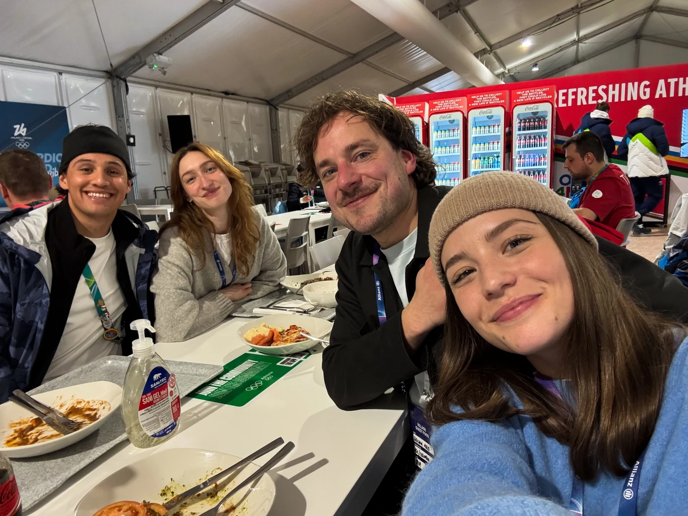 Czwórka uśmiechniętych osób siedzi przy stole podczas posiłku w jasnym namiocie, w tle regały z napojami oraz inni uczestnicy wydarzenia.