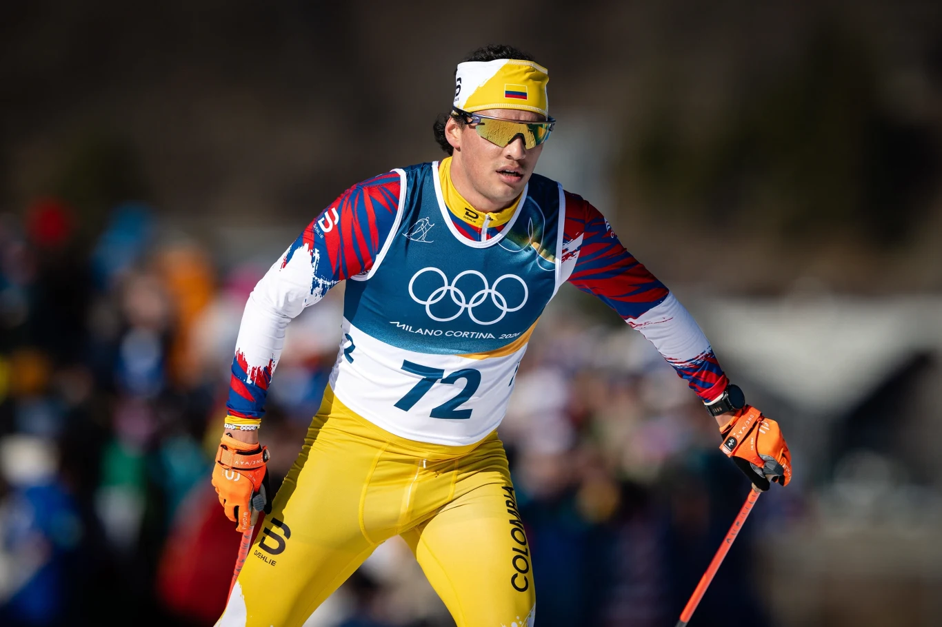 Narciarz biorący udział w wyścigu olimpijskim, ubrany w żółto-niebieski strój z numerem 72, z opaską na głowie i okularami sportowymi, trzyma kijki narciarskie, a w tle rozmazany tłum kibiców.