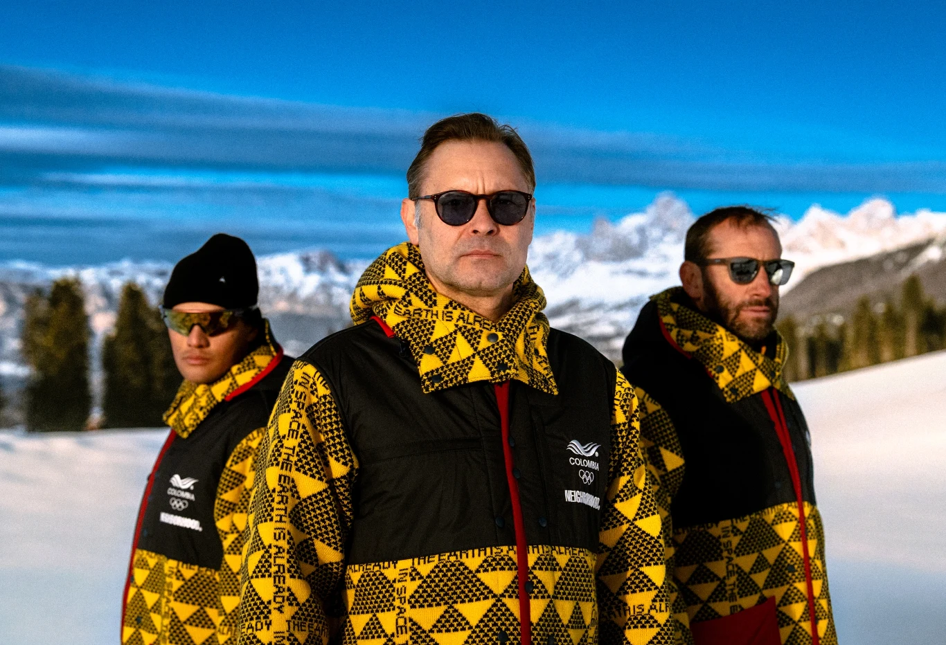 Trzech mężczyzn w jednakowych kurtkach sportowych z geometrycznym wzorem i okularach przeciwsłonecznych stoi na tle ośnieżonych gór i niebieskiego nieba.
