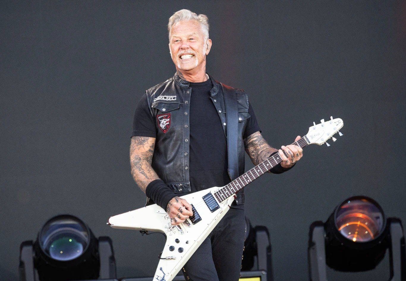 James Hetfield (Metallica) w akcji Mężczyzna grający na białej gitarze elektrycznej na scenie, ubrany w czarną kamizelkę i koszulkę z widocznymi tatuażami na rękach, w tle widoczne reflektory sceniczne.