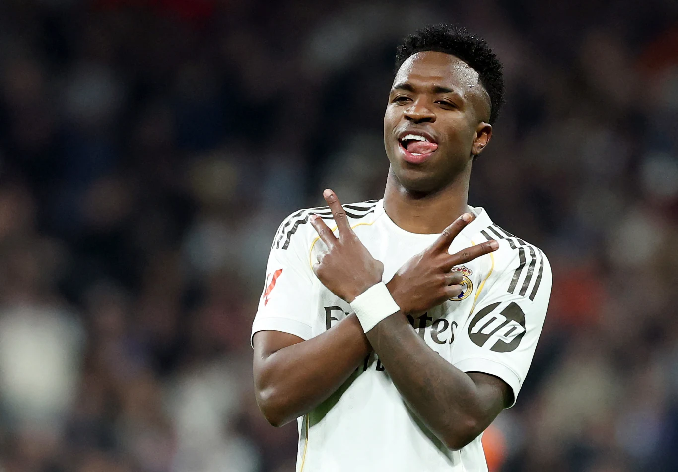 Młody piłkarz w białym stroju sportowym prezentuje gest rękami, uśmiechając się i wystawiając język, w tle rozmyta publiczność na stadionie.