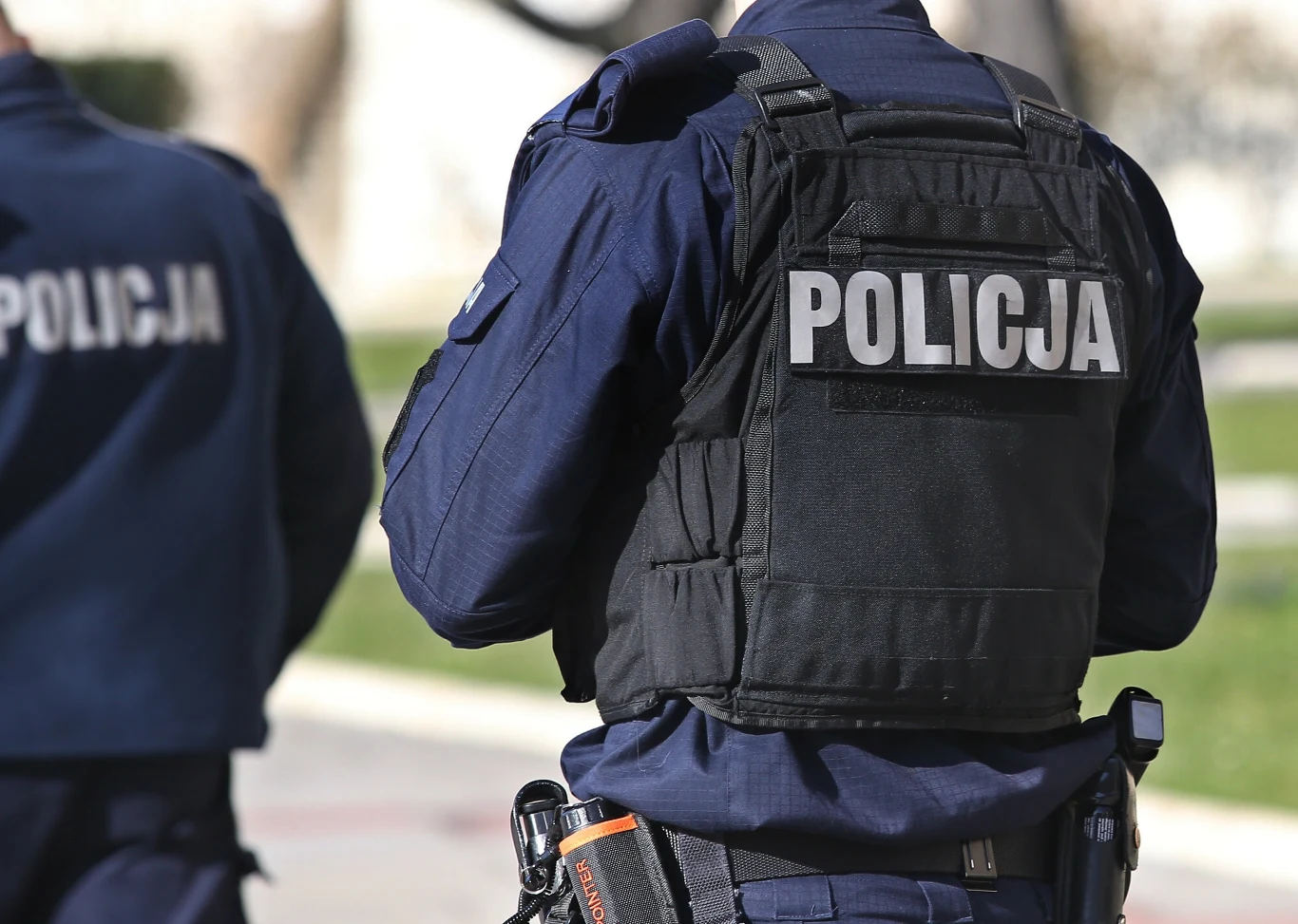 Policjant miał wykorzystać seksualnie funkcjonariuszkę, Mężczyzna został zatrzymany. Zdj. ilustracyjne Dwóch umundurowanych policjantów w kamizelkach taktycznych z widocznym napisem 'Policja' na plecach, stojących na zewnątrz w jasnym świetle, w tle rozmyte przestrzenie miejskie.