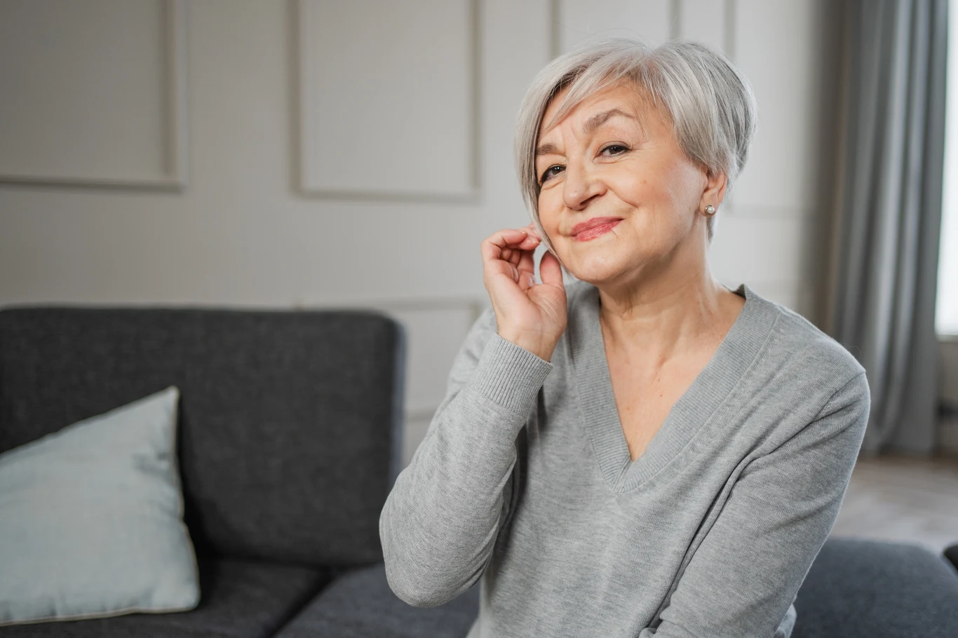 Odpowiednio dobrana grzywka odejmie nawet 10 lat Starsza kobieta o siwych włosach uśmiecha się delikatnie, siedząc na kanapie w jasnym, nowoczesnym wnętrzu. Ubrana jest w szary sweter, jedna ręka spoczywa przy uchu, a na twarzy widoczny jest subtelny makijaż.