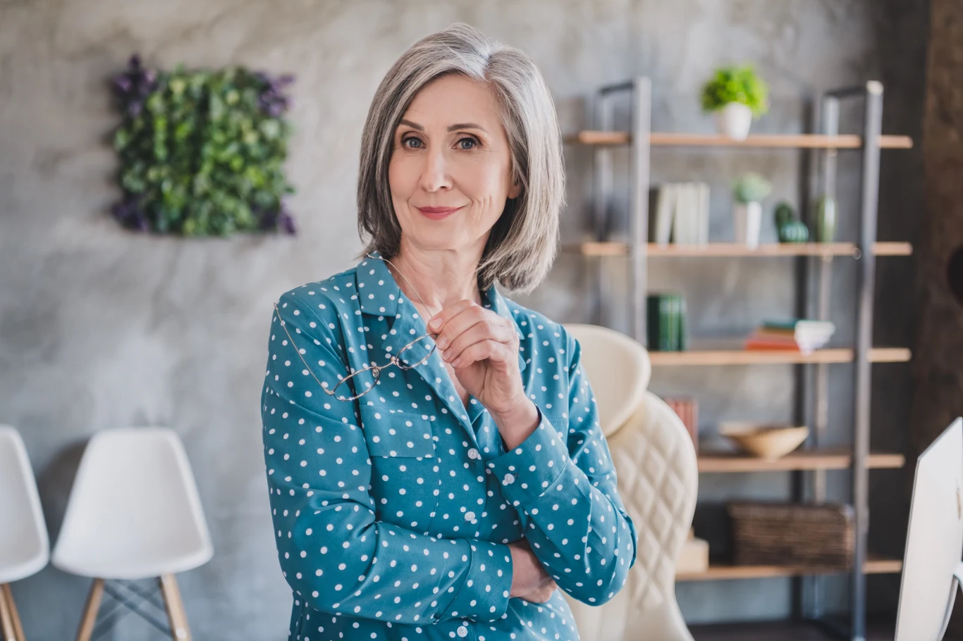 Cieniowane końcówki są hitem fryzjerskim w 2026 Elegancka starsza kobieta z siwymi włosami, ubrana w niebieską koszulę w białe grochy, trzymająca okulary w dłoni, stoi w nowoczesnym, dobrze oświetlonym wnętrzu z regałem i roślinami w tle.