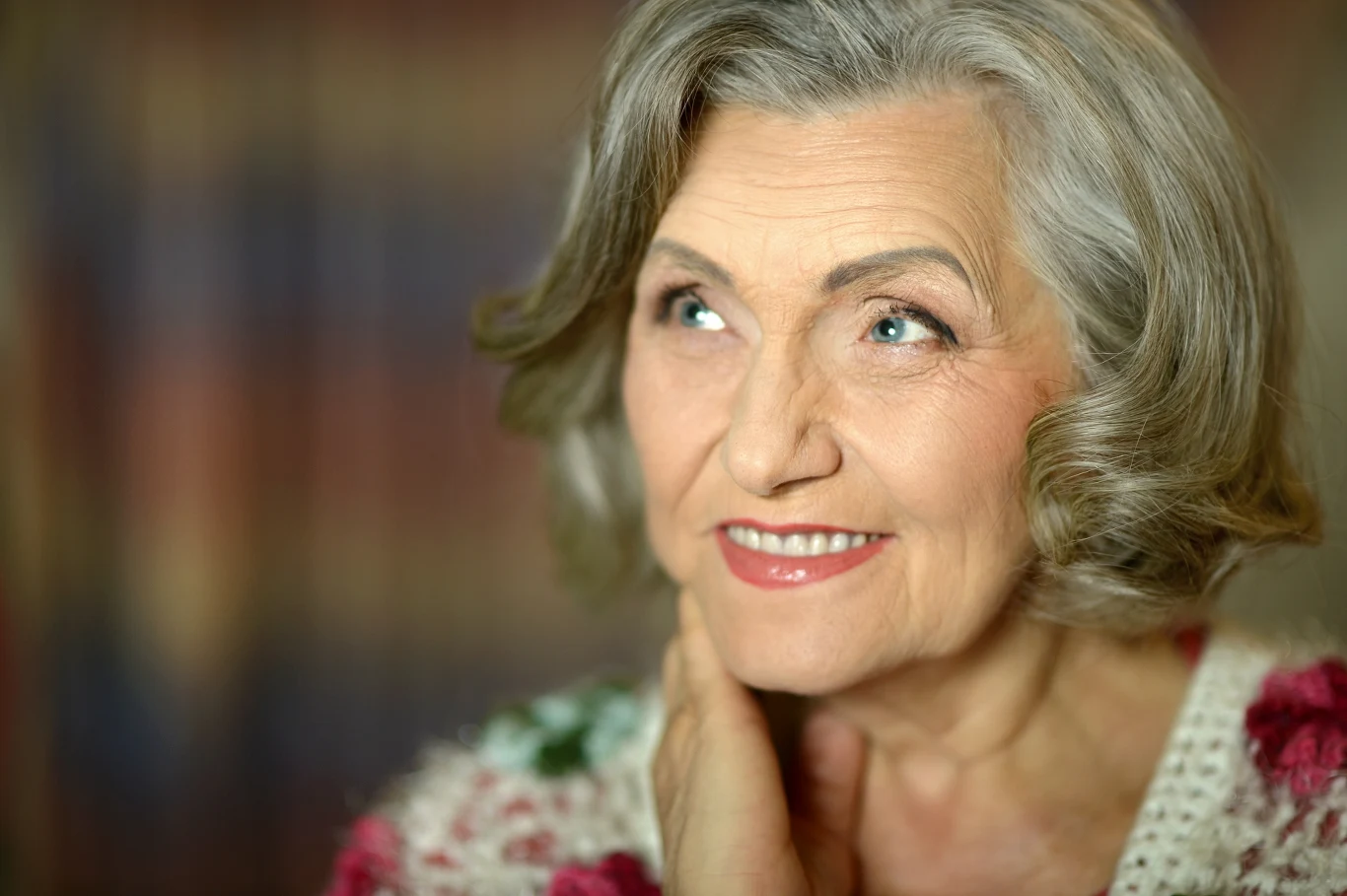 Nie bój się użyć lokówki. Tak odświeżysz swój wygląd Starsza kobieta z siwymi włosami i delikatnym makijażem uśmiecha się lekko, opierając dłoń na policzku. Jej wyraz twarzy jest pogodny, a wzrok skierowany lekko w bok. W tle znajduje się rozmyta kolorowa przestrzeń.