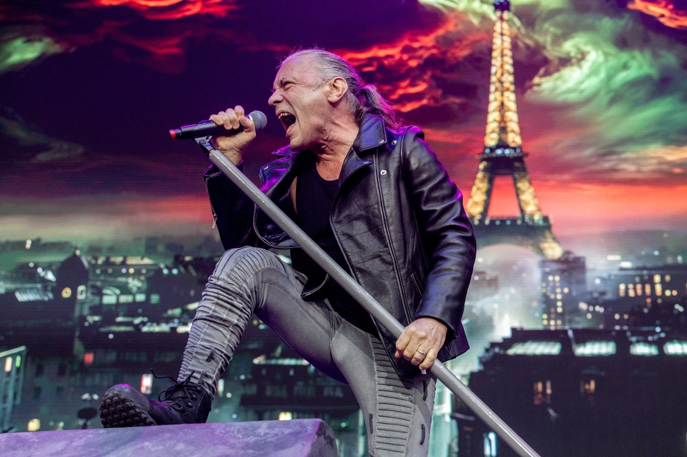 Bruce Dickinson (Iron Maiden) w akcji Wokalista w skórzanej kurtce dynamicznie śpiewa do mikrofonu na tle efektownej scenografii z widokiem na wieżę Eiffla i intensywnie kolorowym niebem.