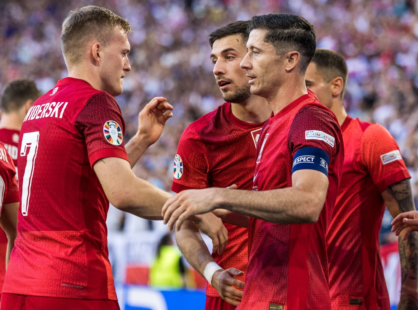 Karol Świderski, Jakub Moder, Robert Lewandowski Grupa piłkarzy w czerwonych koszulkach podczas meczu, skoncentrowani na rozmowie i gratulacjach, w tle rozmyta widownia stadionu.