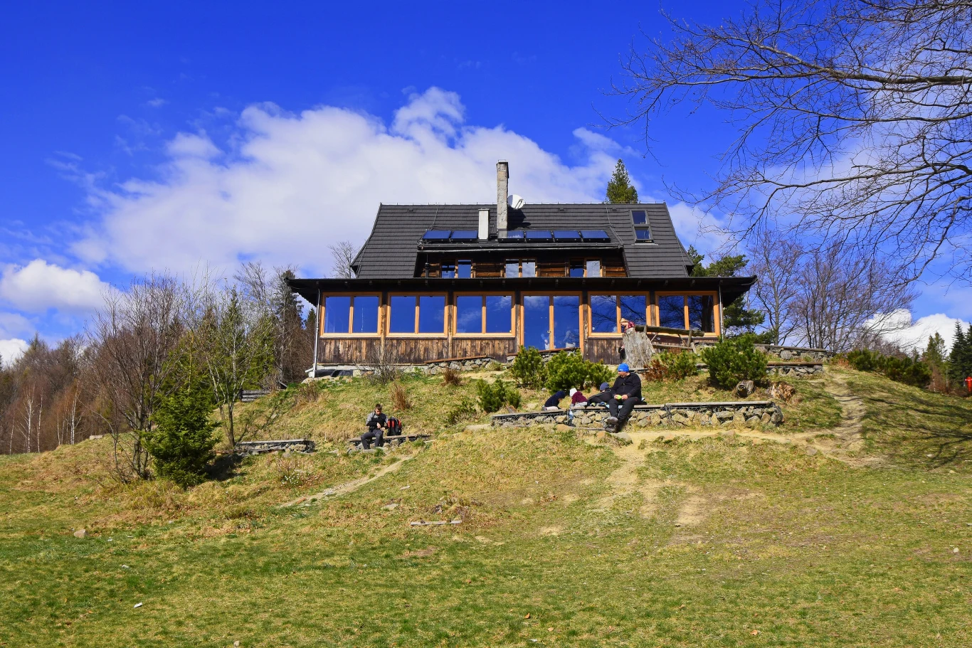 Drewniany schronisko położone na łagodnym wzniesieniu z widokiem na polanę, wokół którego grupka ludzi odpoczywa i korzysta ze słonecznej pogody. Dach budynku wyposażony w kolektory słoneczne, a otoczenie to drzewa i niebieskie niebo z chmurami.