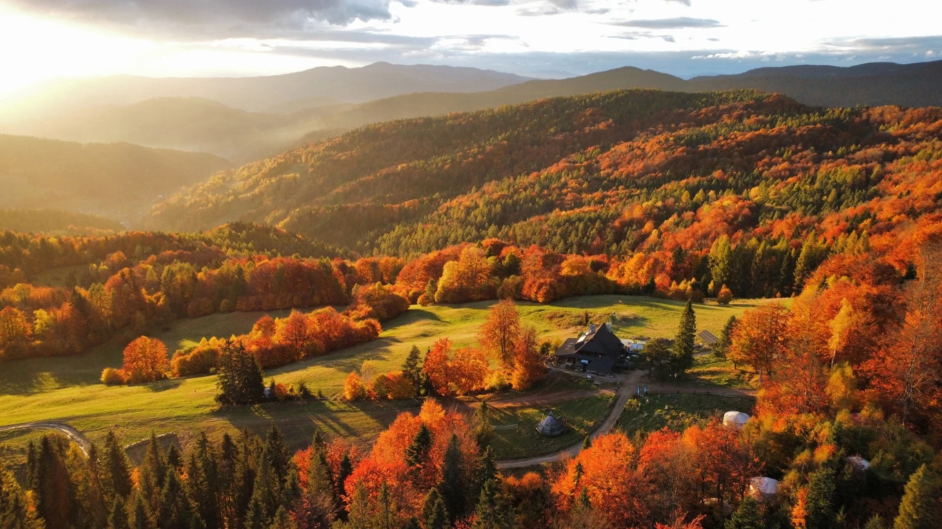 Malownicza dolina otoczona lasami w odcieniach jesieni, pośród których znajduje się samotny dom, a w oddali rozciągają się górzyste wzgórza oświetlone promieniami zachodzącego słońca.
