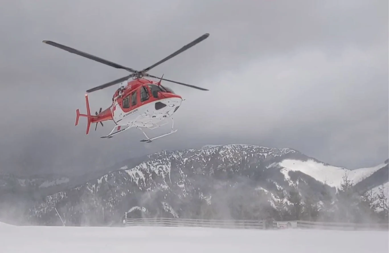Akcja ratunkowa w słowackich tatrach. Zginęło dwóch Polaków. Zdjęcie ilustracyjne Czerwono-biały helikopter unoszący się nad zaśnieżonym terenem górskim, wzbijający śnieg w powietrze, w tle ośnieżone szczyty gór i pochmurne niebo.