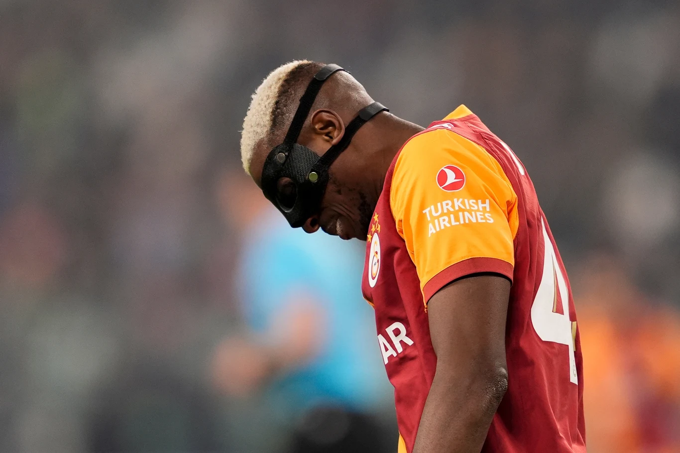 Piłkarz w czerwono-żółtej koszulce Galatasaray z opaską ochronną na twarzy, pochylony głową w dole na tle rozmytego stadionu.