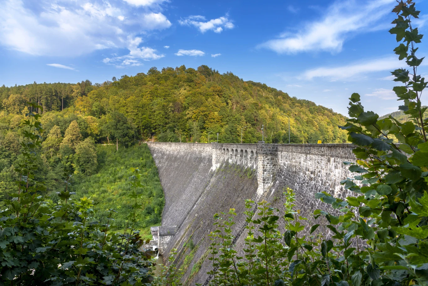 Zapora na Bystrzycy Kamienna zapora wodna otoczona zielenią, tło stanowi gęsty las na wzgórzu pod błękitnym niebem z chmurami.