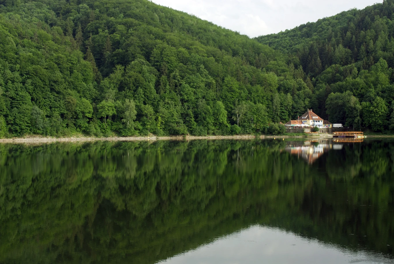 Jezioro Bystrzyckie Jezioro otoczone gęstym lasem, na brzegu widać budynek oraz drewniany pomost, całość odbija się w spokojnej tafli wody, tworząc efekt lustrzanego odbicia.
