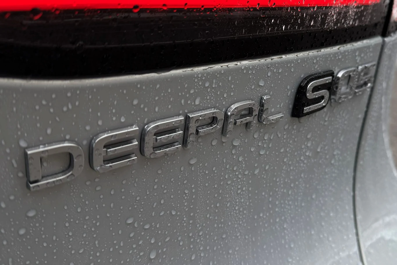 Changan Deepal S05 dostępny jest w Europie z 272-konną wersją RWD i z odmianą AWD, z umieszczonym na przedniej osi 163-konnym silnikiem. Zbliżenie tylnej części srebrnego samochodu marki Deepal S, pokrytego kroplami deszczu oraz z widocznym czerwonym elementem linii tylnego światła.