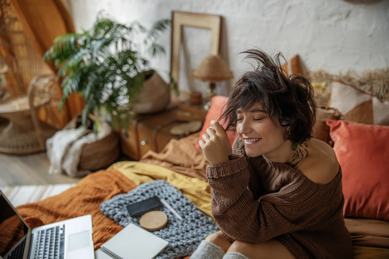 Młoda kobieta z krótkimi, ciemnymi włosami siedzi na przytulnym łóżku w otoczeniu miękkich poduszek i ciepłych tkanin, uśmiechając się z zamkniętymi oczami. Na łóżku obok niej leżą notes, laptop i kawa, a wnętrze urządzone jest w stylu boho z dużą rośl...