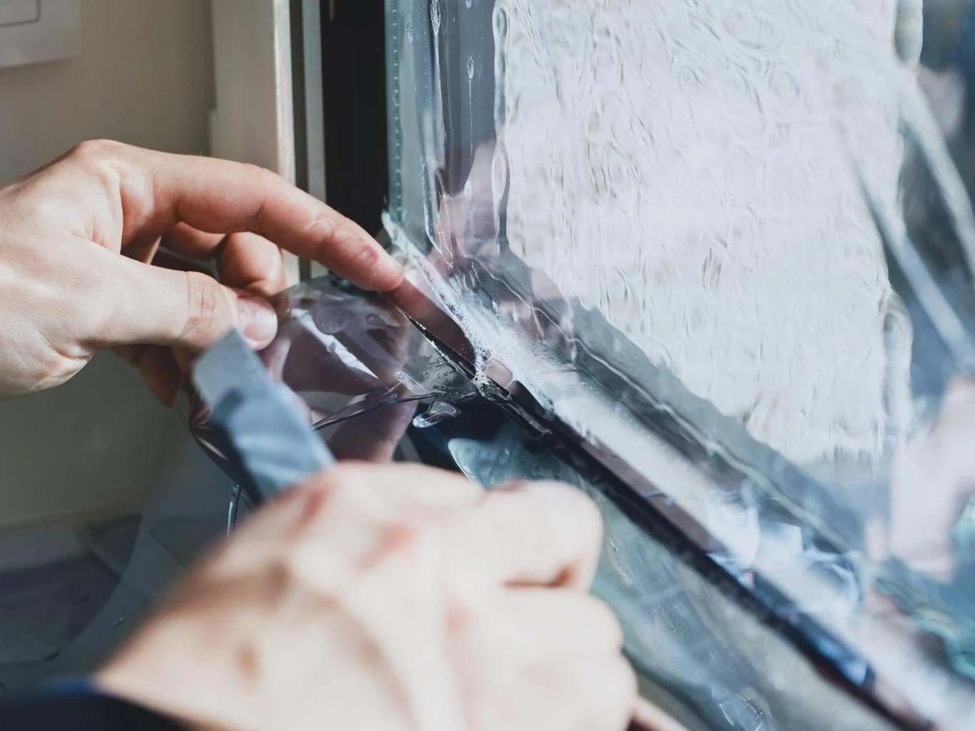 Ręce osoby naklejające przezroczystą folię ochronną na szybę okienną, zbliżenie na szczegółowy moment montażu, widoczna linia łączenia folii z szybą.